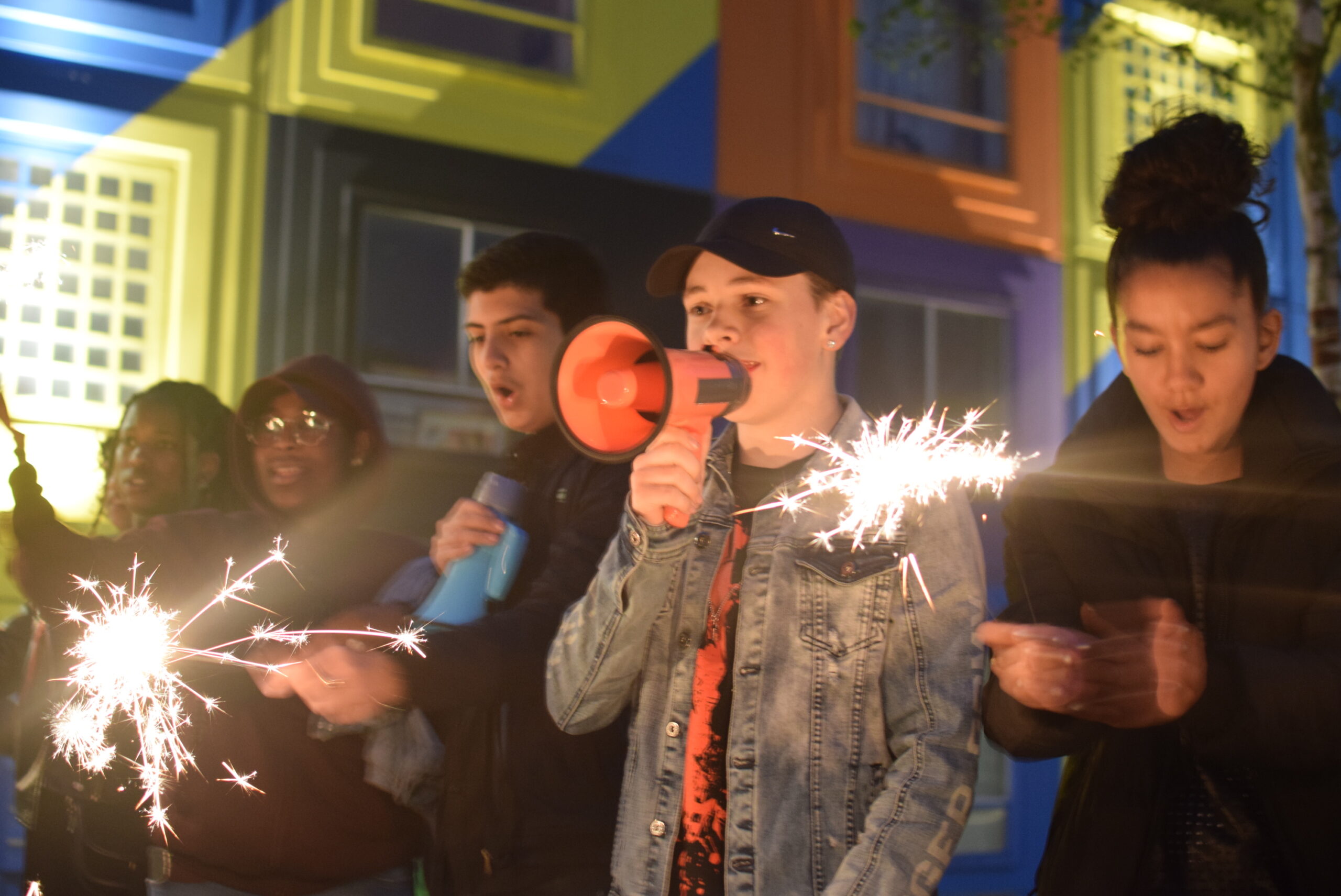 A group of teenagers stands together, some holding fire crackers and one holding a loud speaker.
