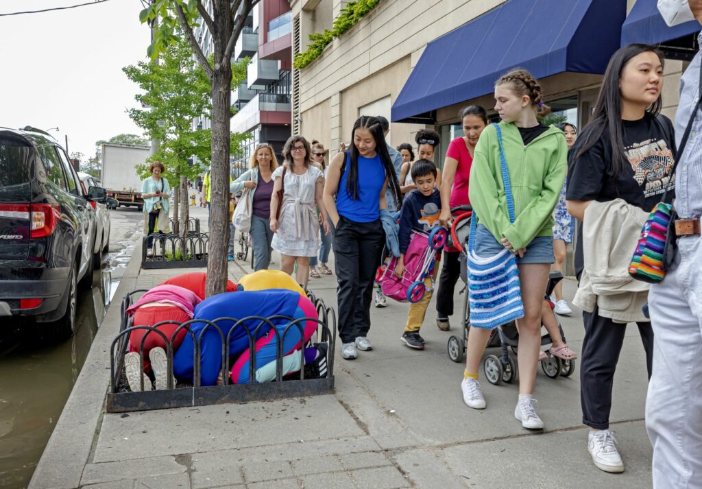 A group of people walk by performers huddled around the base of a tree.